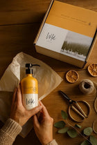Wild England single bottle gift box with model holding sweet orange and cardamom 475ml natural hand and body lotion bottle next to dried oranges, a ball of string and scissors on a rustic wooden surface